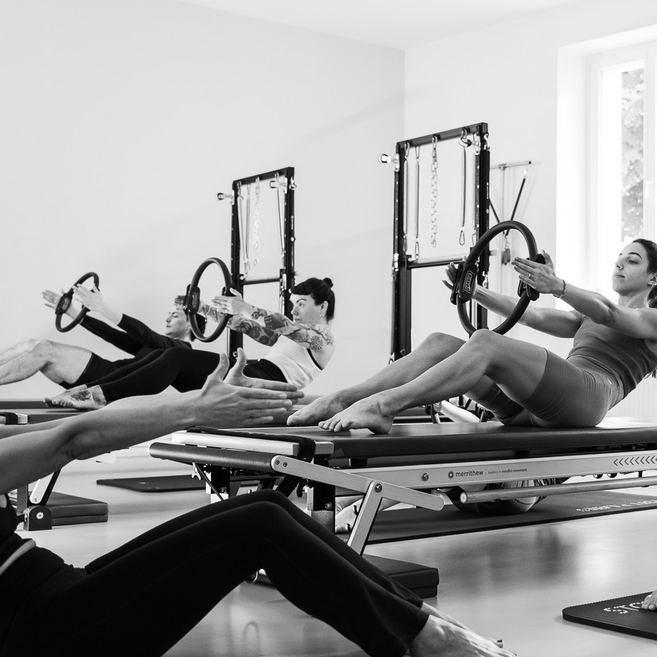 A group is training their core muscles in a Pilates Tower Unit class. They hold the Magic Circle extended in their hands. A Pilates instructor sits in front of them, performing the exercise herself while guiding the group.