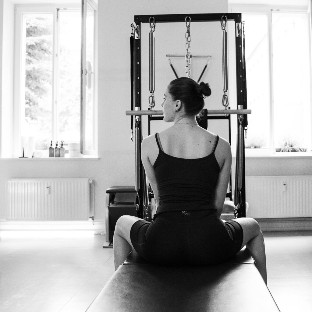 Eine junge Frau sitzt am Pilates-Gerät, der Tower Unit Wall. aufgerichtet, mit dem Blick zum Gerät im Pilates Studio, REALZ Pilates Berlin.