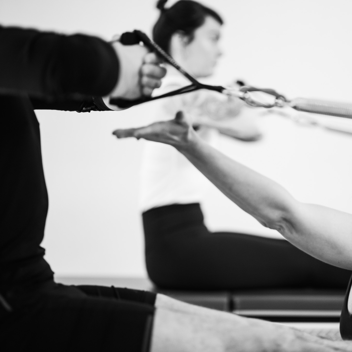 Two people are sitting with fully upright posture at the Tower Unit Wall, holding the hand straps in front of their chest, legs extended. The Pilates instructor indicates the proper height and corrects their body alignment.