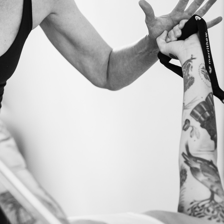 Two arms reach upward holding the Pilates straps from the Tower Unit Wall. A Pilates teacher motivates and guides the person to perform the exercise with correct form.