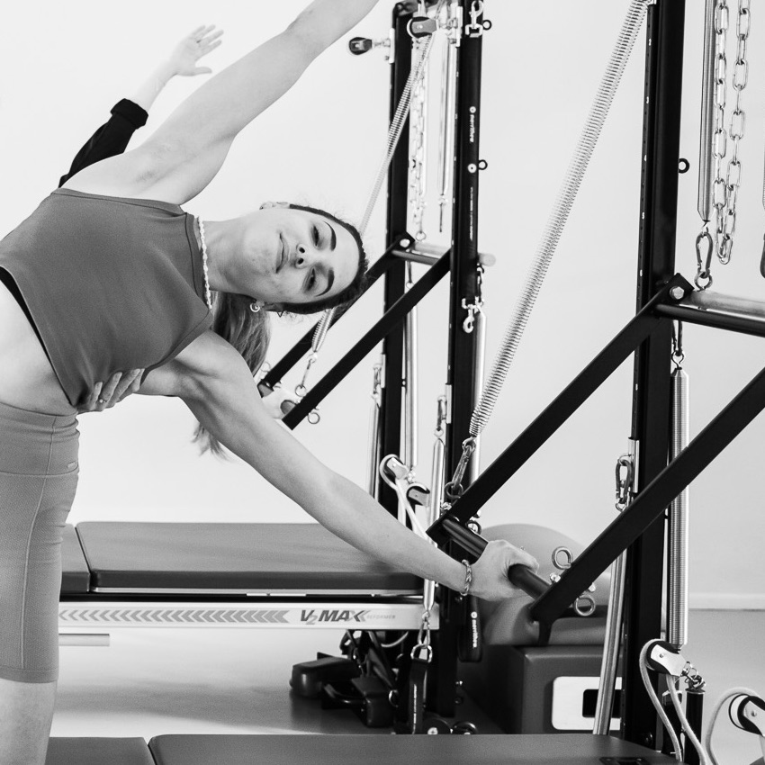 Eine junge Frau trainiert ganz glücklich und ausgeglichen an der Pilates Tower Unit Wall. Sie genießt die Stunde und dehnt sich zur Seite auf, im Pilates Side-Bend Stretch. 