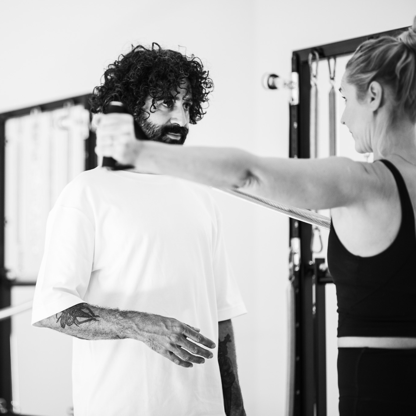 Pilates Solo Training, featuring a young, sporty man at the Tower Unit Wall. The Pilates instructor demonstrates how to correctly extend his arm, and he listens attentively to perform the exercise properly.