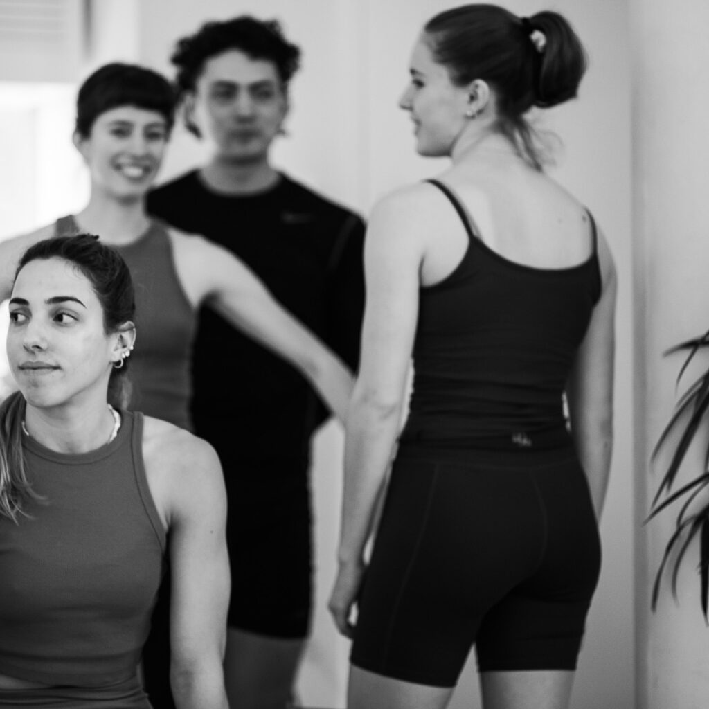 Eine Gruppe junger Pilates Teilnehmer im Studio, die sich vor ihrer Pilates Klasse unterhalten und austauschen. 