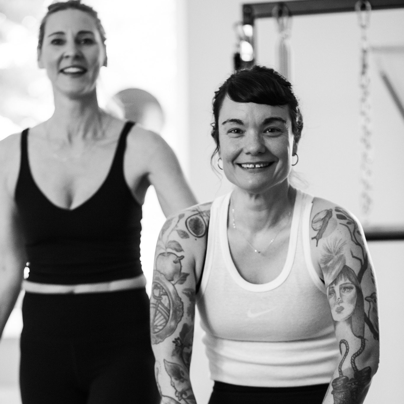 Eine Pilates Teilnehmerin, sitzt ganz glücklich auf dem Pilates Tower Gerät, neben ihr, ihre Pilates-Lehrerin. Beide lächeln, REALZ Pilates Studio Berlin Mitte.