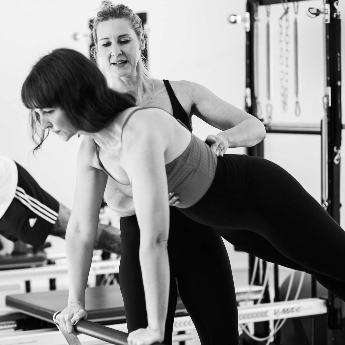 Eine junge Frau trainiert die Plank im Pilates Reformer Training. Kraftvoll drückt sie ihren Oberkörper raus, ihr gesamter Körper ist angehoben, die Arme gestreckt. Eine Pilates Lehrerin steht neben ihr und korrigiert sie in ihrer Rumpfausrichtung.