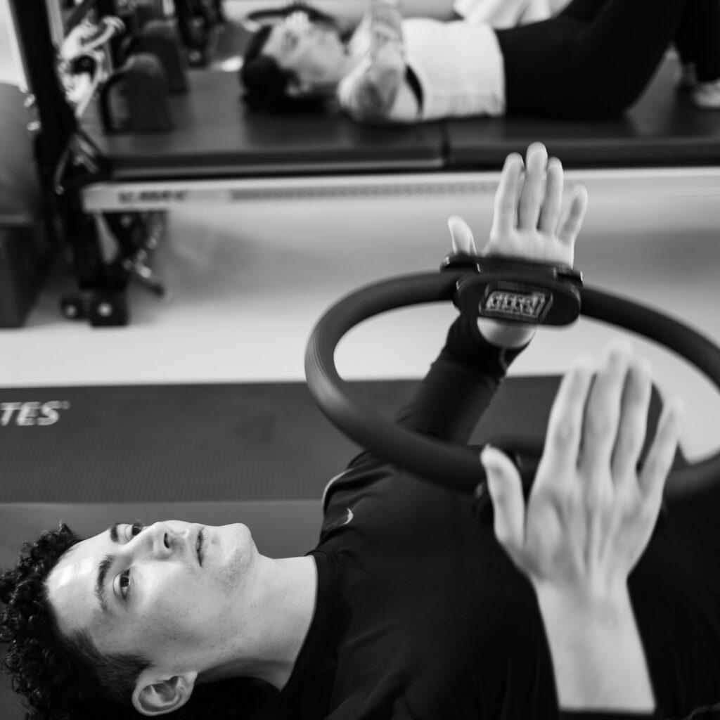 Ein junger Man trainiert in einer Pilates Gruppenklasse, Mattentraining. Er hält dabei ganz konzentriert und mit Kraft den Magic Circle in seinen Händen. Vor seiner Brust und drückt ihn zusammen. Eine Frau ist im Hintergrund mit ihm zu sehen.