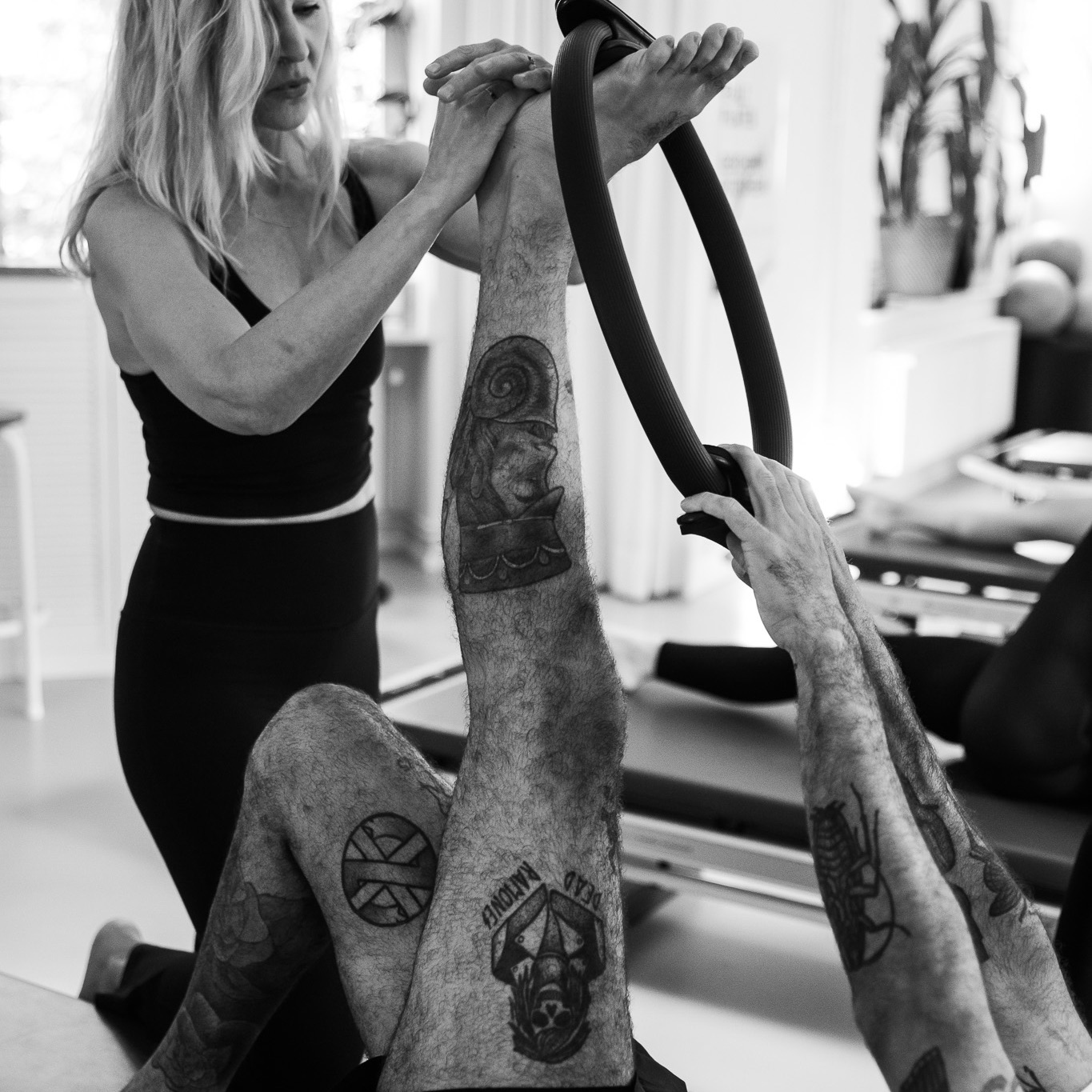 Pilates Gruppenklasse in der Pilates Tower Unit Klasse. Ein Mann dehnt sein Bein mit dem Magic Circle, eine Pilates Lehrerin steht neben ihm und hilft ihn tiefer in die Dehnung zu gehen.