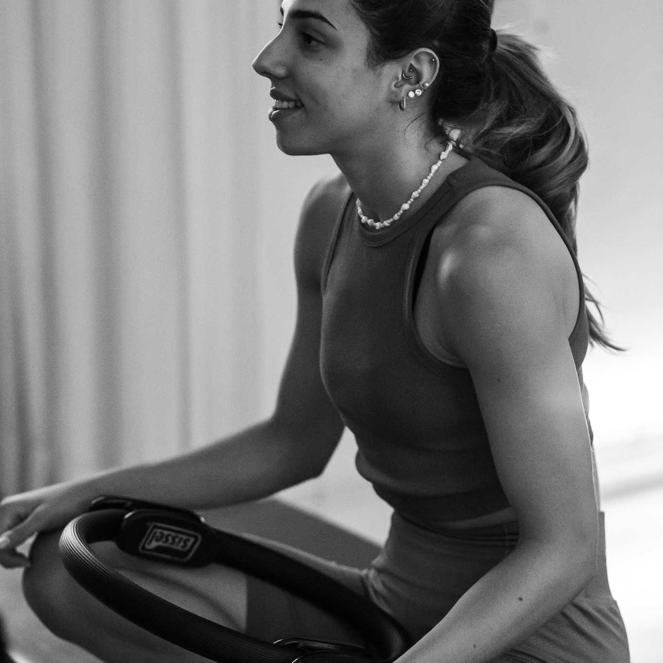 A young woman is sitting on a Pilates machine with a Magic Circle resting on her legs. She looks comfortable, smiling, and completely at ease in the studio.