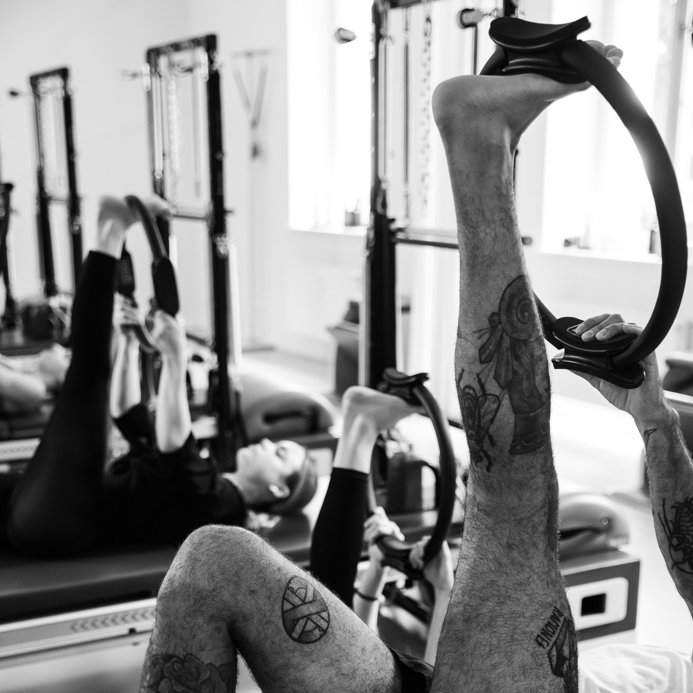Männer und Frauen nehmen am Pilates Workshop teil. Jeder liegt auf seinem Gerät auf der Tower Unit Wall, mit dem Magic Circle am Fuß um den tiefen Stretch zu genießen. Sie halten den Magic Circle mit einer Hand fest um noch tiefer in den Stretch zu gehen und das Bein zu dehnen.