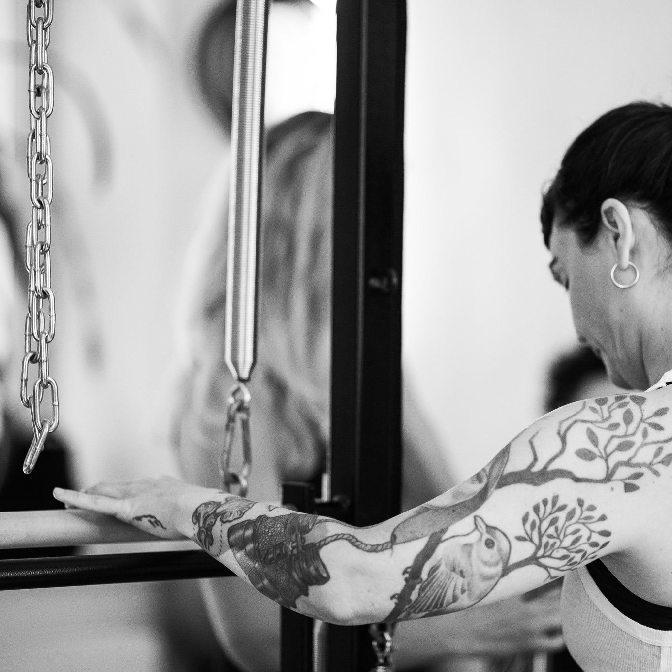 A young woman stands at the Pilates Tower Unit Wall. One hand rests on the barrel, her gaze is soft, and she appears completely centered and grounded. In the background, other people can be seen in the Pilates studio.